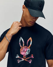 A man in a black Psycho Bunny MENS NOVAN GRAPHIC TEE (MT0200177)—a Pima cotton shirt featuring a vibrant floral bunny skull-and-crossbones logo—looks down, touching his collar with one hand.