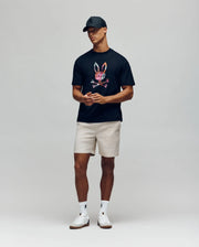 A man in a black cap, beige shorts, white socks and sneakers wears the Psycho Bunny MENS NOVAN GRAPHIC TEE (MT0200177), featuring a colorful bunny print, standing against a plain light gray background.