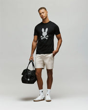 A man wears the Psycho Bunny MENS TALON GRAPHIC TEE (MT0200179), a regular fit black 100% cotton tee with a graphic bunny skull, paired with light shorts, white socks, white sneakers, and holds a black bag in his left hand.