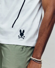 Wearing the Psycho Bunny MENS SOREN GRAPHIC TEE (MT0200180), a 100% cotton white tee with a black bunny and crossbones logo near the hem, and light green shorts, they rest their bracelet-adorned arm along the shirt’s side seam.
