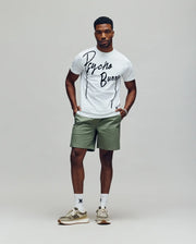 A man stands against a plain background wearing the Psycho Bunny MENS SOREN GRAPHIC TEE (MT0200180) in white, paired with light green shorts, white socks, and beige sneakers. His hands are in his pockets as he looks straight ahead.