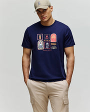 A man wears a beige cap and Psycho Bunny MENS SUNDEW GRAPHIC TEE (MT0200195), a navy Pima cotton shirt with colorful Beach Bunny and palm tree patches, paired with light beige cargo pants, standing against a plain background.