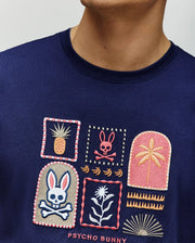 A man wears the Psycho Bunny MENS SUNDEW GRAPHIC TEE (MT0200195), a navy Pima cotton shirt with playful embroidered motifs—pineapple, crossbone rabbit, palm tree, flower, bananas—and "Psycho Bunny" text for a vibrant ocean-inspired style.
