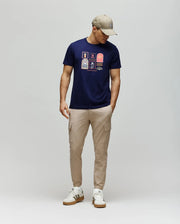 A man wears the Psycho Bunny MENS SUNDEW GRAPHIC TEE (MT0200195) in navy, paired with beige cargo pants, white sneakers, and a beige cap. He stands against a light gray background, looking down with his hands in his pockets.