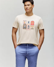 A man stands with his hands in his pockets, wearing the Psycho Bunny MENS SUNDEW GRAPHIC TEE (MT0200195) in beige with a colorful Beach Bunny print and lavender drawstring shorts, looking slightly to the side against a plain background.