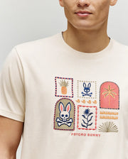 A person wears the Psycho Bunny MENS SUNDEW GRAPHIC TEE (MT0200195), a cream Pima cotton shirt featuring colorful embroidered patches—a bunny with crossbones, pineapple, flower, palm—and "Psycho Bunny" text for an ocean-view vibe.