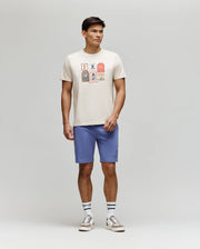 A man stands against a plain background wearing the Psycho Bunny MENS SUNDEW GRAPHIC TEE - MT0200195, light blue shorts, striped white socks, and beige sneakers for an effortless ocean view style.