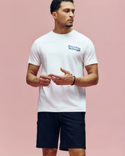 A man stands against a pink background, wearing the Psycho Bunny MENS ARCHIVE WORDMARK GRAPHIC TEE (MT0200030) in white Pima cotton and black shorts. He looks to the side with his hands loosely clasped in front of him.