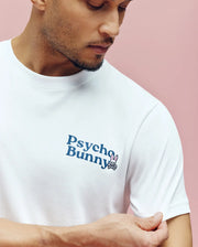 A man wearing the Psycho Bunny MENS ARCHIVE WORDMARK GRAPHIC TEE (MT0200030), a regular fit white Pima cotton shirt with a water-based logo and chest bunny graphic, stands against a pink background and looks down.