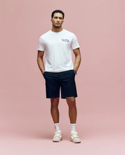 A man stands against a pink background wearing the Psycho Bunny MENS ARCHIVE WORDMARK GRAPHIC TEE (MT0200030), black shorts, white crew socks, and white sneakers, with his hands in his pockets and looking straight ahead.