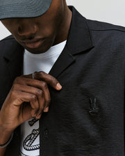 A person wears the Psycho Bunny MENS JAYME KNIT SEERSUCKER CABANA COLLAR SHIRT - MT0300002 with its signature embroidered bunny logo, layered over a white printed tee, paired with a gray cap and ring, gently holding the shirt collar. Their face is partly visible.