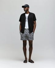A man stands against a plain background wearing the Psycho Bunny MENS JAYME KNIT SEERSUCKER CABANA COLLAR SHIRT - MT0300002 over a white graphic tee, patterned shorts, and black slide sandals. His hands are in his pockets.