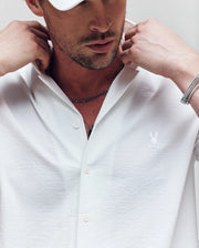 A man adjusts the cabana collar of his white Psycho Bunny JAYME KNIT SEERSUCKER shirt (MT0300002) with an embroidered bunny logo. He also wears a silver chain, bracelet, and white cap in a closely cropped upper body shot.