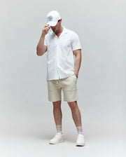 A man stands against a plain background wearing the Psycho Bunny MENS JAYME KNIT SEERSUCKER CABANA COLLAR SHIRT (MT0300002), paired with beige shorts, white sneakers, socks, and a white cap. He looks down, touching the brim of his hat.