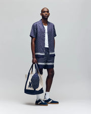 A man wears the Psycho Bunny MENS HUNTER TENCEL SHIRT - MT0300016, paired with matching shorts, a white tank, navy sneakers with white socks, and carries a beige tote bag featuring a navy design, all set against a plain background.