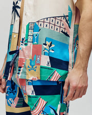 A person wears a partially buttoned, tucked-in Psycho Bunny MENS BLAISE TENCEL SHIRT (MT0300056) and matching shorts, both in breathable Tencel with colorful abstract beach-themed patterns, palm trees, and Psycho Bunny logos.