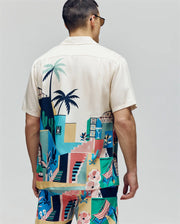 A man is seen from the back wearing Psycho Bunny's MENS BLAISE TENCEL SHIRT - MT0300056, a breathable short-sleeve shirt with a colorful tropical graphic of stairs, plants, and buildings. He also wears matching shorts and sunglasses.