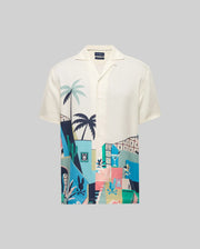 The Psycho Bunny Mens Blaise Tencel Shirt (MT0300056) is a cream, short-sleeved top with breathable fabric and a vibrant abstract tropical print of palm trees, buildings, and playful animals in blue, green, pink, and yellow.