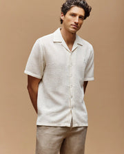 A man with wavy brown hair wears the Psycho Bunny MENS SAMMY CROCHET SHORT SLEEVE SHIRT (MT0300108) in cream and light brown pants, standing with his hands behind his back against a neutral beige background.