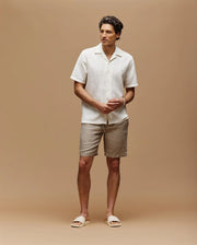Against a beige background, a man with short dark hair wears the Psycho Bunny MENS SAMMY CROCHET SHORT SLEEVE SHIRT (MT0300108) in white, paired with light tan shorts and beige slide sandals. His relaxed posture features hands loosely clasped.