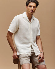 A man sits on a wooden stool against a beige background, wearing the Psycho Bunny MENS SAMMY CROCHET SHORT SLEEVE SHIRT (MT0300108) in cream and light tan shorts, looking at the camera with a neutral expression, one hand resting on his knee.