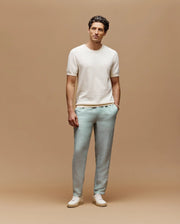 A man stands against a tan background wearing the Psycho Bunny MENS MARIO STRIPED SWEATER T-SHIRT (MT0400009), light blue pants, and white sneakers, with one hand in his pocket and a relaxed expression.