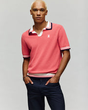 A man with short, bleached blond hair models the Psycho Bunny MENS CARTER JOHNNY COLLAR KNIT POLO - MT0400093, a 100% cotton pink polo with white and navy trim, paired with dark pants against a plain light background.