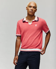 A man with short bleached hair wears the Psycho Bunny MENS CARTER JOHNNY COLLAR KNIT POLO (MT0400093) in coral, featuring white and navy trim, paired with navy pants, standing against a plain light gray background.