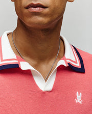 A close-up shows a person wearing the Psycho Bunny MENS CARTER JOHNNY COLLAR KNIT POLO (MT0400093) in pink 100% cotton, featuring white and navy trim, a white rabbit logo, and a silver chain necklace. Only the lower face and neck are visible.