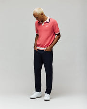 A person with short blond hair wears the Psycho Bunny MENS CARTER JOHNNY COLLAR KNIT POLO (MT0400093) in pink, paired with dark pants and white sneakers, standing against a plain light gray background and looking down with hands in pockets.