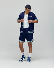 A man stands against a plain background wearing the Psycho Bunny MENS BASIL TAPED CAMP COLLAR SHIRT (MT0500004) in a premium cotton-modal blend, styled with matching shorts, a white tank top, white socks, and blue-and-white sneakers. He looks down adjusting his shirt.