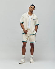 A man stands confidently in the Psycho Bunny MENS BASIL TAPED CAMP COLLAR SHIRT (MT0500004), paired with matching cream shorts with green trim, a white tank, cream sneakers, and patterned white socks against a neutral background.