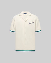 The Psycho Bunny MENS BASIL TAPED CAMP COLLAR SHIRT (MT0500004) is a cream, short-sleeve shirt with a Johnny collar, teal and black striped sleeves and hem, embroidered "Psycho Bunny" on the chest, made from cotton-modal blend. Shown on gray.