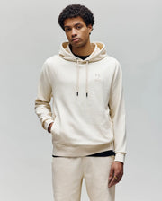 A young man with short curly hair wears the Psycho Bunny MENS KAI TWILL TEXTURE HOODIE (MT0500019) in cream, featuring an embroidered Bunny logo and matching sweatpants, standing against a plain light gray background.