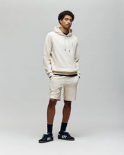 A young man stands facing forward in a light beige Psycho Bunny MENS KAI TWILL TEXTURE HOODIE (MT0500019) with an embroidered Bunny logo, matching shorts, black socks, and black Adidas sneakers against a plain light grey background.
