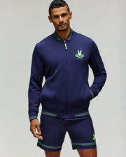 A man wears the Psycho Bunny TENNIS BALL CREW MENS JACKET ENGLISH (MT0500049)—an athletic zip jacket with green trim and embroidered Bunny logo—standing against a plain light gray background.