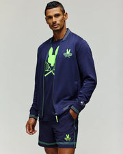 A man stands confidently against a plain light background wearing the Psycho Bunny TENNIS BALL CREW MENS JACKET ENGLISH (MT0500049), matching shorts, and a tennis-themed neon green graphic T-shirt.