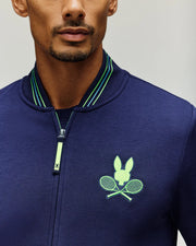 A man wears the Psycho Bunny Tennis Ball Crew Men's Jacket, featuring an embroidered Bunny with green and blue crossed rackets on the left chest and a green-striped collar, highlighting its stretch modal-cotton fabric and signature logo.