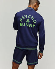 A man stands facing away, wearing the Psycho Bunny TENNIS BALL CREW MENS JACKET ENGLISH - MT0500049. The stretch modal-cotton jacket’s back displays "PSYCHO BUNNY" and an embroidered Bunny skull logo in bright green.
