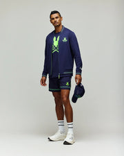 A man poses against a plain background wearing the Psycho Bunny Tennis Ball Crew Mens Jacket (MT0500049) in navy, matching shorts, a neon green graphic tee, white socks and sneakers, holding a navy cap in his left hand.