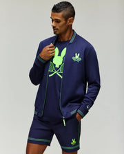 A man wears the Psycho Bunny TENNIS BALL CREW MENS JACKET FRENCH (MT0500050), a navy blue stretch modal-cotton athletic jacket with neon green embroidered Bunny and tennis racket designs, standing against a plain light background.