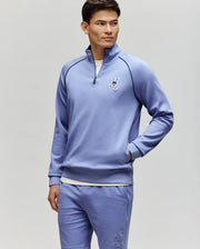 A man wears the Psycho Bunny MENS SHAWN 1/4 ZIP SWEATSHIRT (MT0500115) and matching shorts, both light purple with a white bunny logo, standing with one hand in his pocket against a plain light gray background.