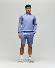 A man stands against a plain background wearing the Psycho Bunny MENS SHAWN 1/4 ZIP SWEATSHIRT (MT0500115) in light purple, with matching shorts, white socks with black stripes, and beige sneakers. His hands are in his pockets and he looks ahead.