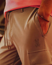 A person wears tan shorts with Psycho Bunny’s embroidered bunny and crossbones logo, a pink shirt, and the MENS LEATHER BRAIDED BRACELET (MO1700020) from Psycho Bunny featuring a hinged clasp on their wrist.