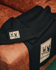 A black cotton-wool blend beanie and luxury Mens Monroe Twill Patch Scarf from Psycho Bunny, featuring white embroidered logos of a stylized bunny and skull crossbones, laid on a brown leather surface.