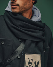 A person in a luxury coat with large buttons wears the Psycho Bunny MENS MONROE TWILL PATCH SCARF, a black cotton wool blend with a rabbit motif. They sport a gray hoodie and carry a leather strap with partially visible text against a green background.