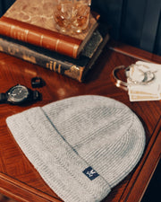 A MENS RACKED BEANIE - B6A188E200 from Psycho Bunny, crafted in a warm gray Merino wool knit, rests on a wooden table. Nearby, there are stacked vintage books, a wristwatch, a glass with a drink, and a set of playing cards.