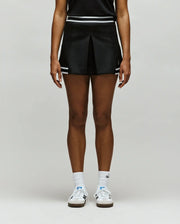 A person stands against a plain background, wearing the Psycho Bunny WOMENS KNIT SKORT - WO1900001 in black with white stripes, paired with a black top, white socks, and white sneakers. The image is cropped from the shoulders down.