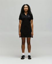 A person with curly hair faces forward, wearing the Psycho Bunny WOMENS POLO DRESS - WO2000003 in black with an embroidered Bunny, paired with white socks and black sneakers, against a plain light gray background.
