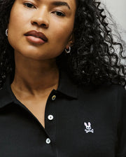 A woman with curly hair wears the Psycho Bunny WOMENS POLO DRESS (WO2000003) in black, featuring an embroidered Bunny logo above white crossbones on the chest. Silver hoop earrings complete her look, and the dress flatters her silhouette.